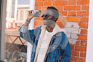 Portrait happy smiling african man with vintage film camera taking picture walking on city street over brick wall