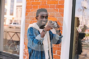 Portrait happy smiling african man with vintage film camera taking picture walking on city street over brick wall