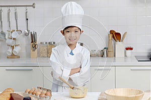 Portrait of happy smiling Asian boy in white chef uniform with hat standing with arms crossed and mixing eggs bowl on table at