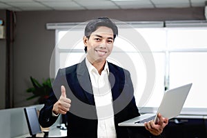 Portrait of happy smiling Asian businessman in black suit giving thumb up while holding laptop computer, standing inside office