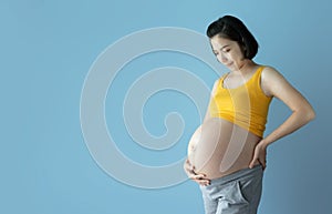 Portrait of Happy Smiling Asian Pregnant woman standing over blue isolated background. Young Female touching her belly while her