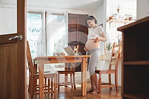 Portrait of Happy Smiling Asian pregnant woman standing showing her belly using video chatting with family on laptop