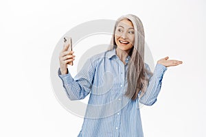 Portrait of happy smiling asian senior woman, old lady showing smth while on video call, talking with mobile phone app