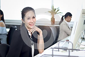 Portrait of happy smiling Asian woman officer sitting at office desk with blurred busy working colleague background, Modern