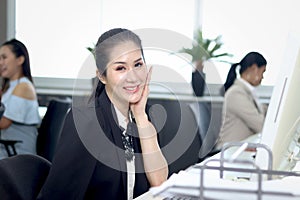 Portrait of happy smiling Asian woman officer sitting at office desk with blurred busy working colleague background, Modern