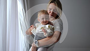 Portrait of happy smiling baby girl laughing looking away in hands of confident beautiful mother looking out the window