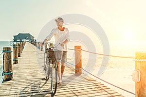 Portrait of a happy smiling barefoot man dressed in light summer clothes and sunglasses riding a bicycle on the wooden sea pier.