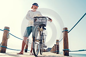 Portrait of a happy smiling barefoot man dressed in light summer clothes and sunglasses riding a bicycle on the wooden sea pier.