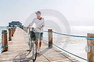 Portrait of a happy smiling barefoot man dressed in light summer clothes and sunglasses walking with a bicycle on the wooden sea