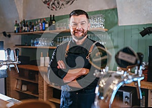Portrait of happy smiling bearded barman dressed in a black uniform with an apron at bar counter with draught beer taps.