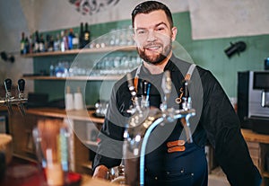 Portrait of happy smiling bearded barman dressed in a black uniform with an apron at bar counter with draught beer taps.