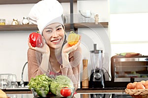 Portrait of happy smiling beautiful Asian woman wearing apron and chef hat holding bell peppers at kitchen with fresh vegetables