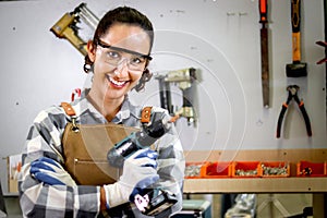 Portrait of happy smiling beautiful carpenter woman wearing safety glasses goggles and apron holding hand drill, female craft