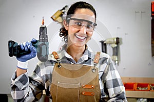 Portrait of happy smiling beautiful carpenter woman wearing safety glasses goggles and apron holding hand drill, female craft