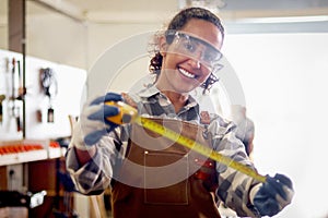 Portrait of happy smiling beautiful carpenter woman wearing safety glasses goggles and apron holding yellow tape measure, female
