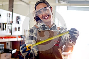 Portrait of happy smiling beautiful carpenter woman wearing safety glasses goggles and apron holding yellow tape measure, female
