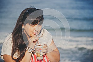 Portrait of happy smiling beautiful overweight young woman in white T-shirt drinking sweet coffee through straw outdoors at beach