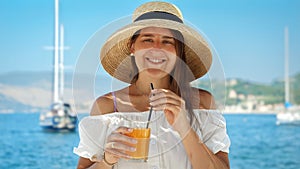 Portrait of happy smiling brunette woman drinking orange juice against sea beach view and moored yachts