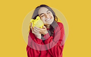 Portrait of happy smiling brunette woman snuggling up to yellow piggy bank on yellow background.