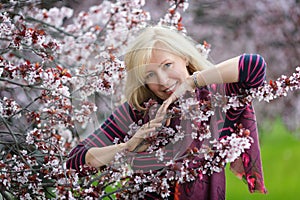 Portrait of happy smiling Caucasian blond woman with long hair near blossoming plum cherry tree, no teeth, looking to the camera