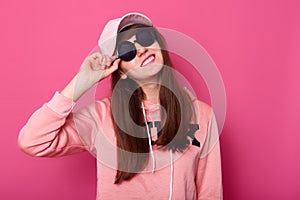 Portrait of happy smiling Caucasian brunette teenager in stylish short pink hoodie, rose cap, black sunglasses, holds hads on hat