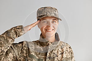 Portrait of happy smiling Caucasian woman army soldier saluting while posing against light background indoor, looking at camera