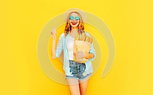 Portrait happy smiling cool girl holding paper bag with long white bread baguette, wearing straw hat, shorts on colorful yellow