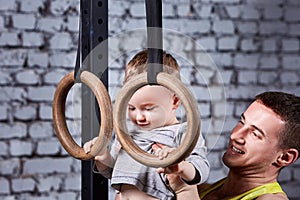 Portrait of happy smiling father and little son exercising with gimnastic rings against brick wall in the gym.