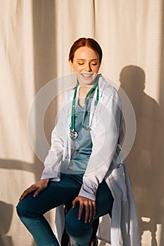 Portrait of happy smiling female doctor in white coat sitting on chair near window in sunny day in medical clinic office