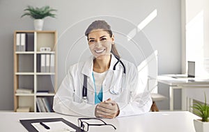 Portrait of happy smiling female doctor in white coat sitting at desk in her office