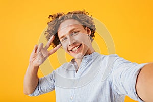 Portrait of happy smiling guy 20s taking selfie photo and showing peace sign, isolated over yellow background