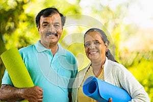 portrait of Happy smiling indian senior couple with yoga mat looking at camera during morning workout at park - concept