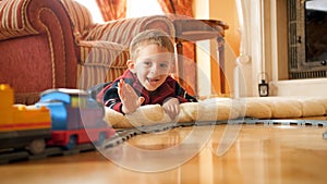 Portrait of happy smiling little boy lying on floor and waving hand to toy train riding on railroad at living room.