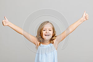 Portrait of a happy smiling little child girl blonde raised her hands up on a gray background. Success concept