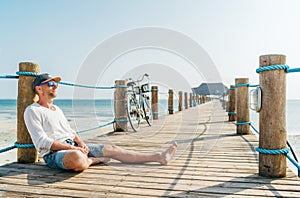 Portrait of a happy smiling man dressed in light summer clothes and sunglasses sitting and enjoying time on wooden sea pier.