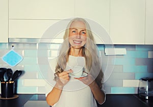 Portrait of happy smiling middle-aged gray-haired woman enjoying hot tasty cup of coffee or tea in the kitchen in early morning at