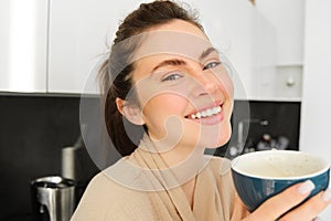 Portrait of happy, smiling modern woman, starts her day with morning mug of tea. Girl drinking coffee in the kitchen