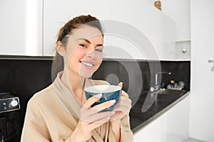 Portrait of happy, smiling modern woman, starts her day with morning mug of tea. Girl drinking coffee in the kitchen