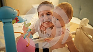 Portrait of happy smiling mother hugging her adorable baby boy in diapers standing in bed at night