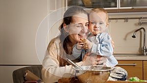 Portrait of happy smiling mother hugging her little baby son covered in dough and flour while baking cake or bread at