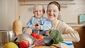 Portrait of happy smiling mother with little baby son having fun on kitchen while cooking. Concept of little chef, children