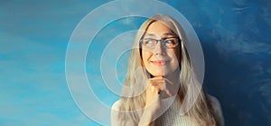 Portrait of happy smiling pensive middle-aged woman in eyeglasses looking away. Gray-haired female thinking on blue background