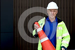 Portrait of happy smiling senior elderly mature engineer wearing safety vest and helmet standing and holding traffic cone, worker