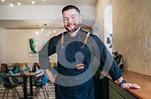Portrait of happy smiling small business owner dressed in a black chef uniform with an apron in his cozy restaurant hall.