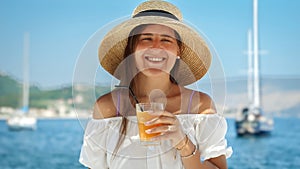 Portrait of happy smiling woman in big hat enjoying her cocktail or fresh juice against sea and yachts view