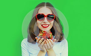 Portrait of happy smiling woman with handful of fresh strawberries wearing a red heart shaped sunglasses on green