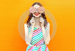 Portrait happy smiling woman hiding her eyes with two lollipop in colorful striped dress on orange wall