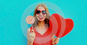 Happy smiling woman holding lollipop and bunch of red heart shaped balloons wearing a sunglasses on a blue background