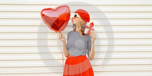Portrait of happy smiling woman with red heart shaped balloon and gift box wearing beret and skirt on white background
