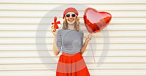 Portrait of happy smiling woman with red heart shaped balloon and gift box wearing beret and skirt on white background